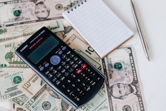 calculator, note pad, and pen on top of dollar bills